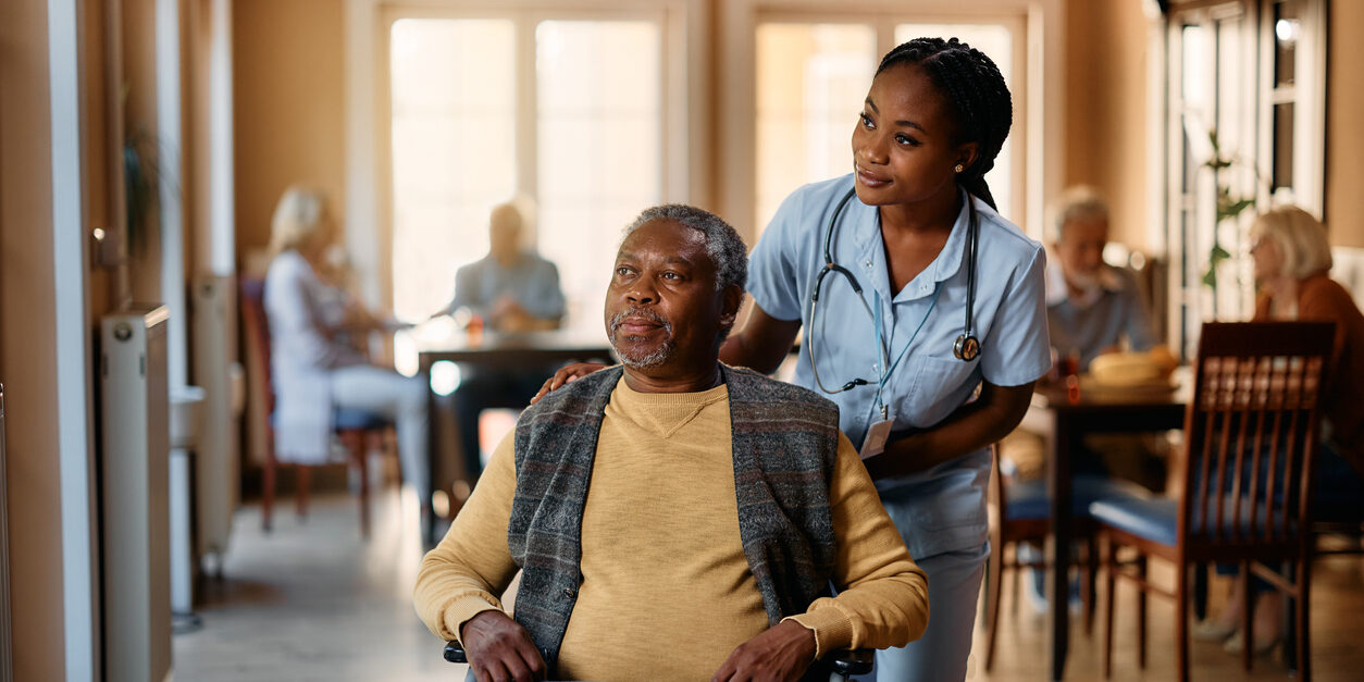 Black nurse and senior man in wheelchair looking through the window at nursing home.