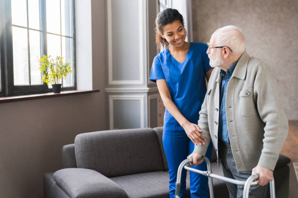 7 Portrait of an african young nurse helping old elderly disable man grandfather to walk using walker equipment in the bedroom. Senior patient of nursing home moving with walking frame and nurse support
